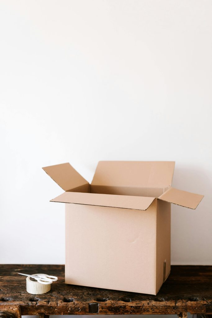 An open cardboard box on a weathered wooden table with tape and scissors beside it, ready for packing.